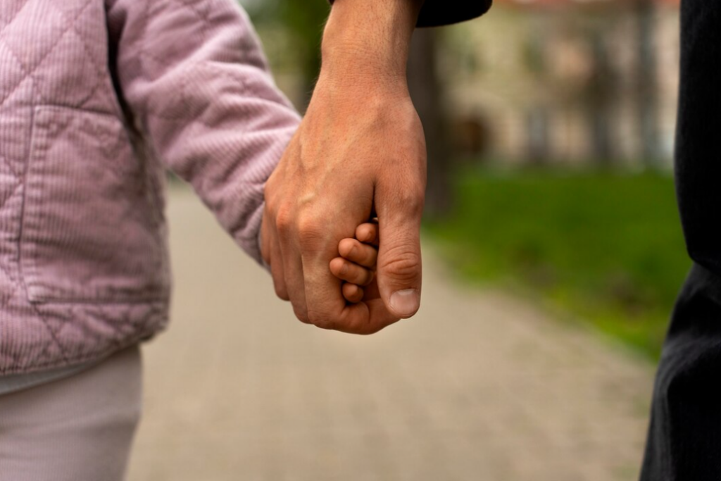 young child and adult holding each other's hands, a blurred road and grass behind